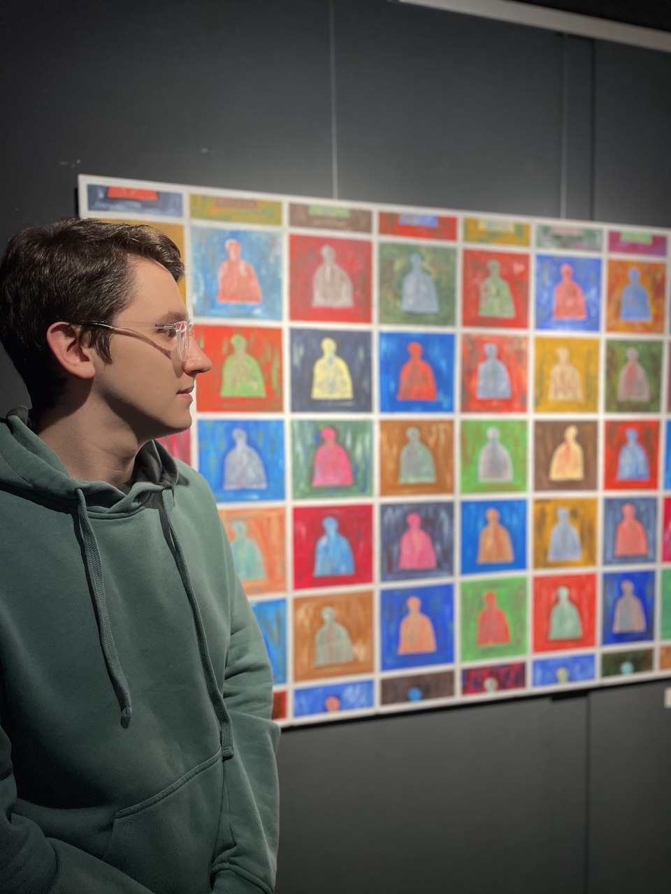 A man with glasses, wearing a teal-colored hoodie, is looking to the side, away from the camera. He is standing indoors in front of a large, colorful artwork composed of a grid of many small, repeating figures on different colored backgrounds.